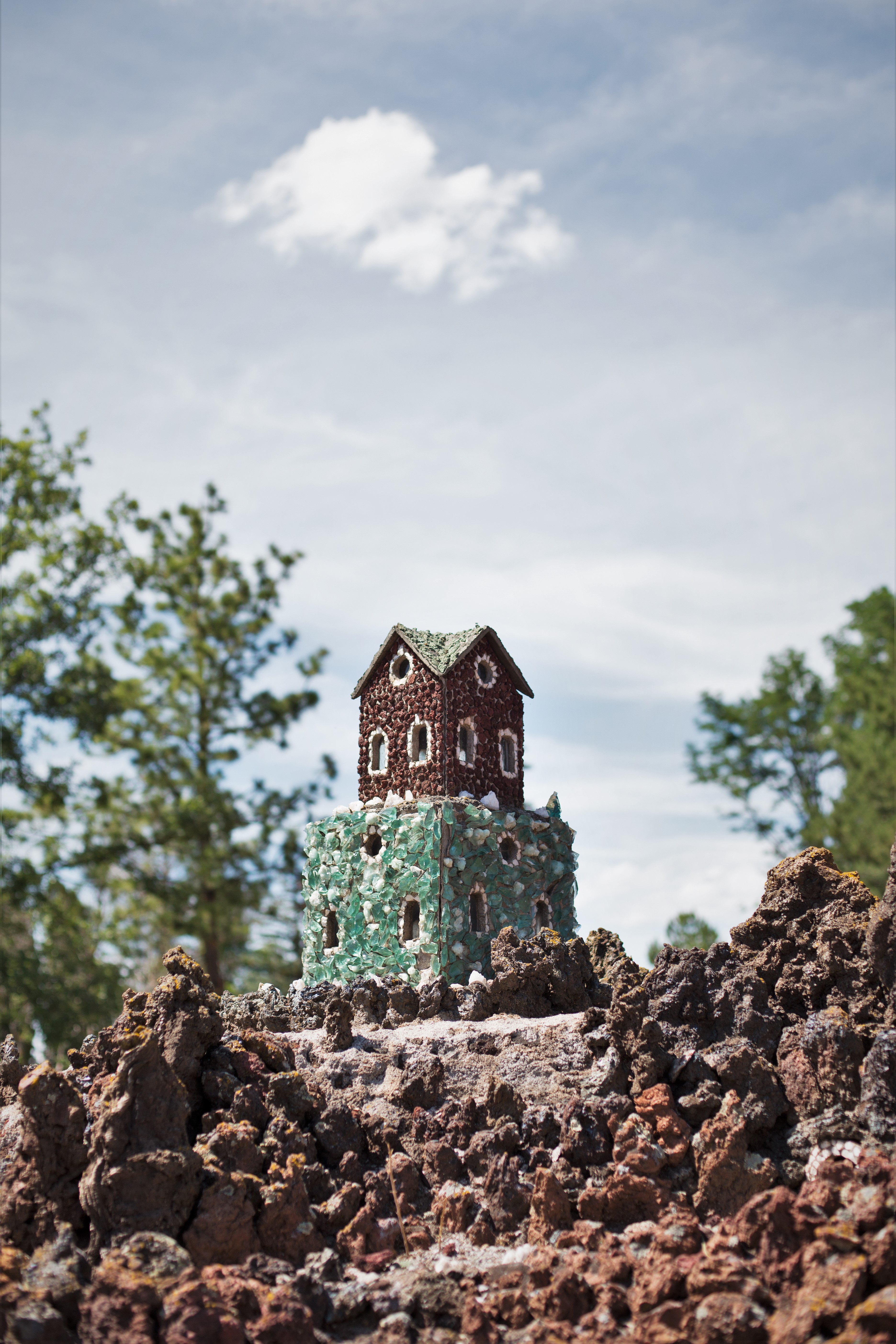 A tiny handmade house on clumps of dirt against a blue, cloudy sky. Represents "The Memory House," a childhood home